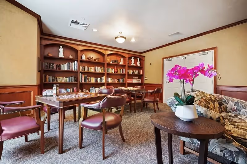 A cozy interior room with a wooden bookshelf filled with books and decorative items along the back wall. In front of the bookshelf are tables with chairs arranged around them, suitable for reading or playing games. To the right, there is a floral-patterned couch next to a round wooden table holding a white vase with vibrant pink orchids. The room has beige walls with wooden paneling and a carpeted floor.