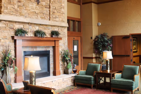 Cozy senior living facility common area with a stone fireplace, wooden mantel, two green upholstered armchairs, side tables with lamps, and decorative plants around the fireplace and in the corner.