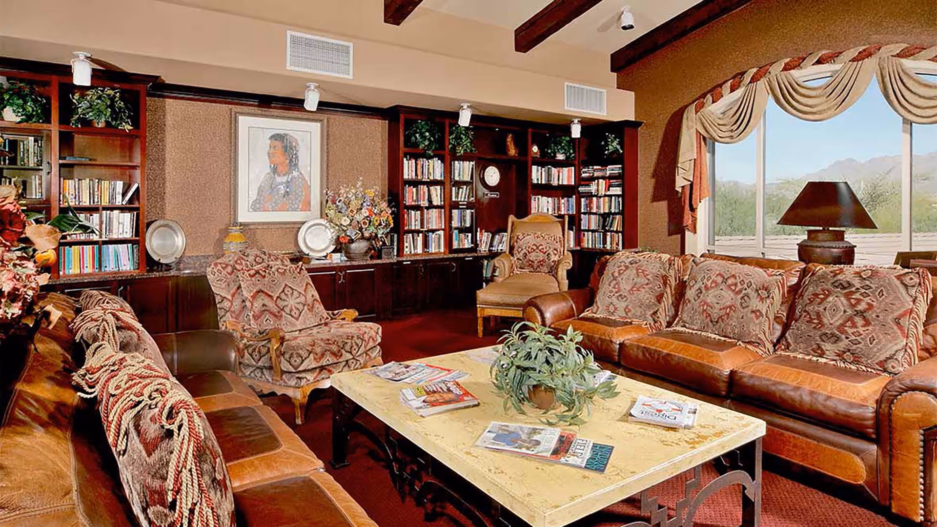 A cozy living room with leather sofas and patterned cushions, a marble coffee table with magazines and a plant, wooden bookshelves filled with books and decorative items, a framed portrait on the wall, and a large window with a view of mountains and greenery outside.