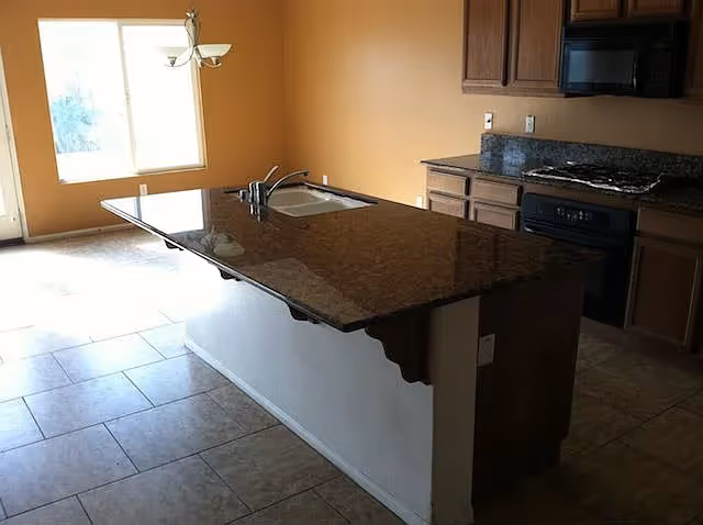 Kitchen interior with a granite-topped island and sink, stove and wooden cabinets on a tiled floor.