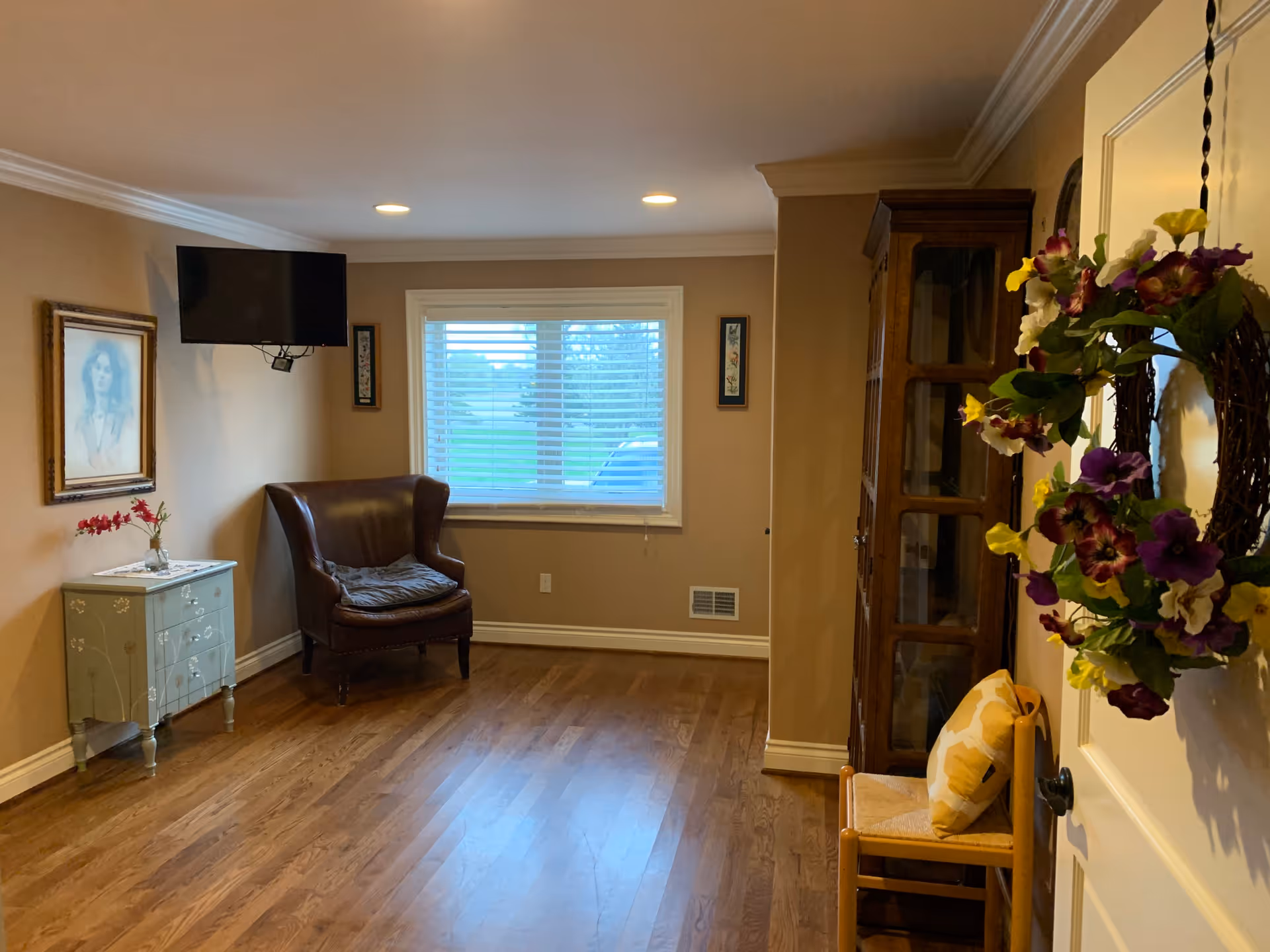 A cozy living room with wooden flooring, beige walls, and a window with white blinds. The room features a brown leather armchair with a cushion, a small decorative cabinet with a vase of flowers, a wall-mounted flat-screen TV, a wooden display cabinet, and a wooden chair with a yellow patterned cushion. A colorful floral wreath hangs on the door.