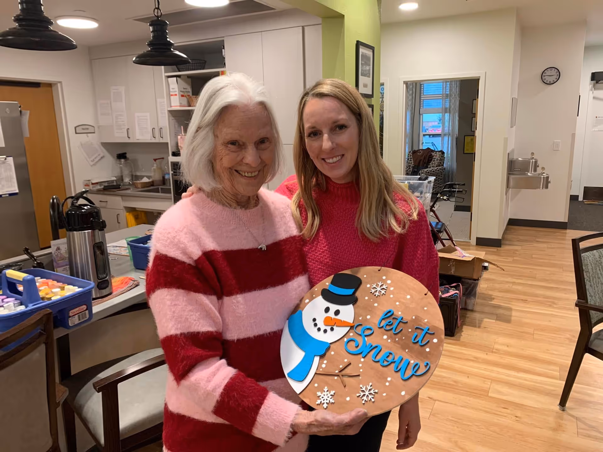 An elderly woman and a younger woman standing together in a kitchen area, smiling at the camera. The elderly woman is wearing a pink and red striped sweater and holding a circular wooden decoration with a snowman and the words 'let it snow'. The kitchen has white cabinets, a coffee dispenser, and various items on the counter. The background shows a hallway and some chairs.