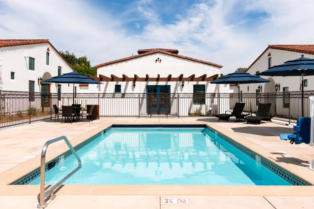 Outdoor swimming pool with lounge chairs, umbrellas, and white Mediterranean-style buildings in the background.