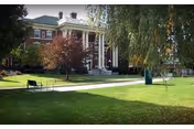 Large historic brick building with white columns at the entrance, surrounded by green lawn, trees, and benches on a sunny day.