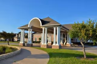 Entrance area of a senior living facility with a covered drop-off zone supported by white columns, surrounded by green grass and trees under a clear blue sky.