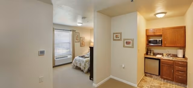 Interior view of a senior living facility apartment showing a small kitchenette with wooden cabinets, a microwave, mini fridge, and sink on the right. To the left, there is a bedroom area with a bed, window with blinds, and an air conditioning unit below the window. The walls are light-colored and there are framed pictures hanging on the walls.