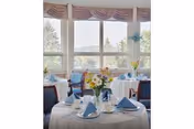 A dining room with round tables covered in white tablecloths, each set with blue folded napkins, glassware, and floral centerpieces. Large windows in the background offer a view of trees and distant hills under a partly cloudy sky.