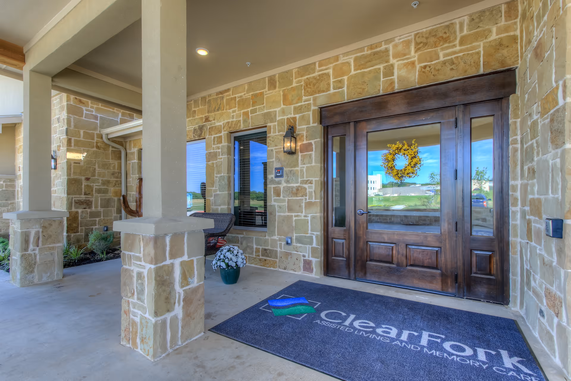 Entrance of Clear Fork Assisted Living and Memory Care facility featuring a stone exterior, wooden double doors with glass panels, a yellow wreath hanging on the door, a welcome mat with the facility's name, two stone pillars, a window with blinds, a wall-mounted lantern light, and a potted plant with white flowers.