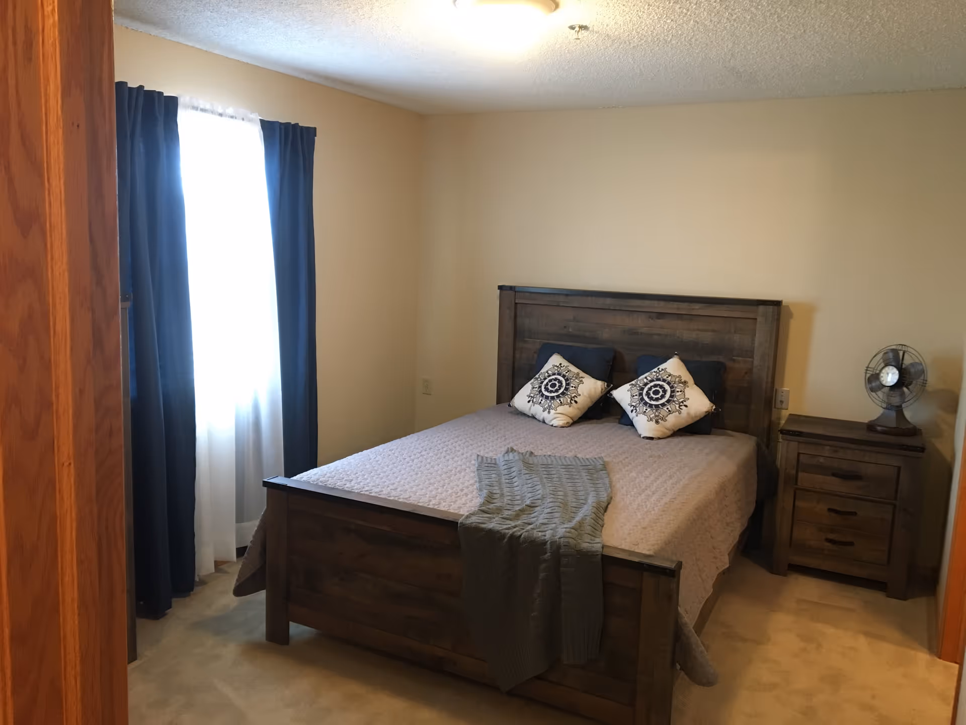 A bedroom with a wooden bed frame and matching nightstand. The bed is made with a light-colored quilt, two decorative pillows, and a folded blanket at the foot. A small vintage-style fan sits on the nightstand. There is a window with dark blue curtains and sheer white curtains behind the bed.