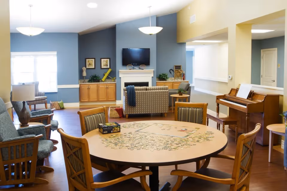 A cozy living room area in Randall Residence of Tipp City featuring a round table with a partially completed jigsaw puzzle and chairs around it. In the background, there are comfortable armchairs, a piano with sheet music, a flat-screen TV mounted above a white fireplace, wooden cabinets, and decorative plants. The walls are painted in blue and beige tones, and the room is well-lit with ceiling lights and natural light from a window.