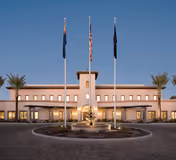 Front exterior view of the Arizona State Veteran Home in Tucson, featuring a symmetrical building with a central tower, three flagpoles with flags, palm trees, and a circular driveway with a fountain in the foreground.