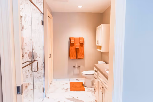 Bright bathroom interior with a glass-enclosed shower, toilet, vanity, and orange towels and bath mat.