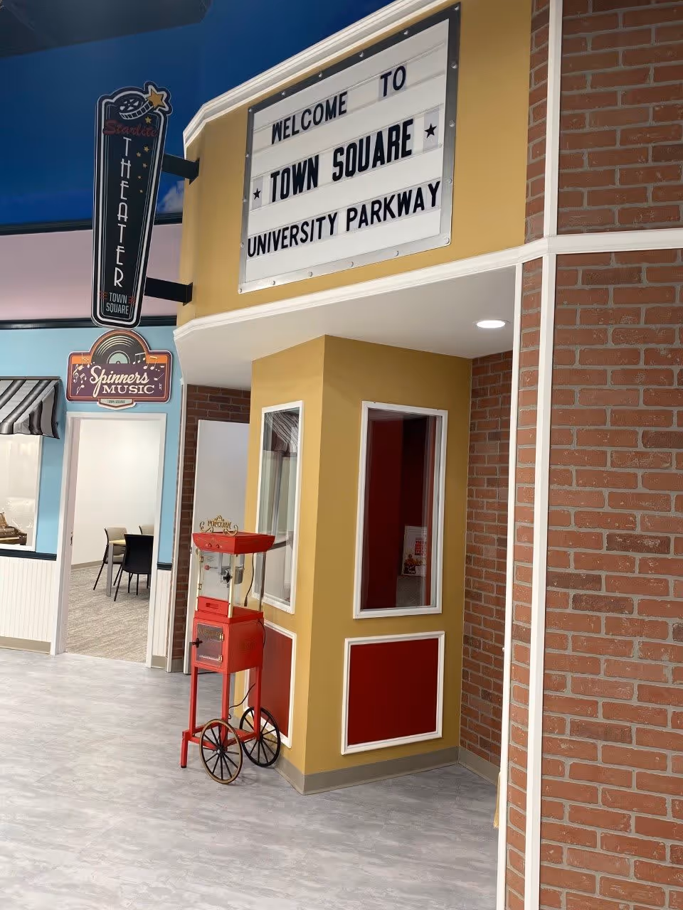 Interior view of a senior living facility area with a vintage-style popcorn machine on wheels, a yellow and red ticket booth, and a sign above that reads 'Welcome to Town Square University Parkway'. To the left, there are signs for 'Starlite Theater' and 'Spinner's Music' above doorways leading to other rooms.