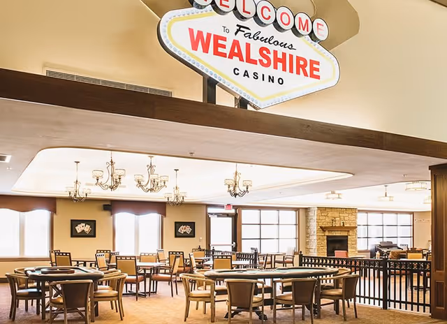 Interior view of a spacious room with multiple tables and chairs arranged for group seating. The room features chandeliers hanging from the ceiling, large windows allowing natural light, and a stone fireplace in the background. A large sign above reads 'Welcome to Fabulous Wealshire Casino'.