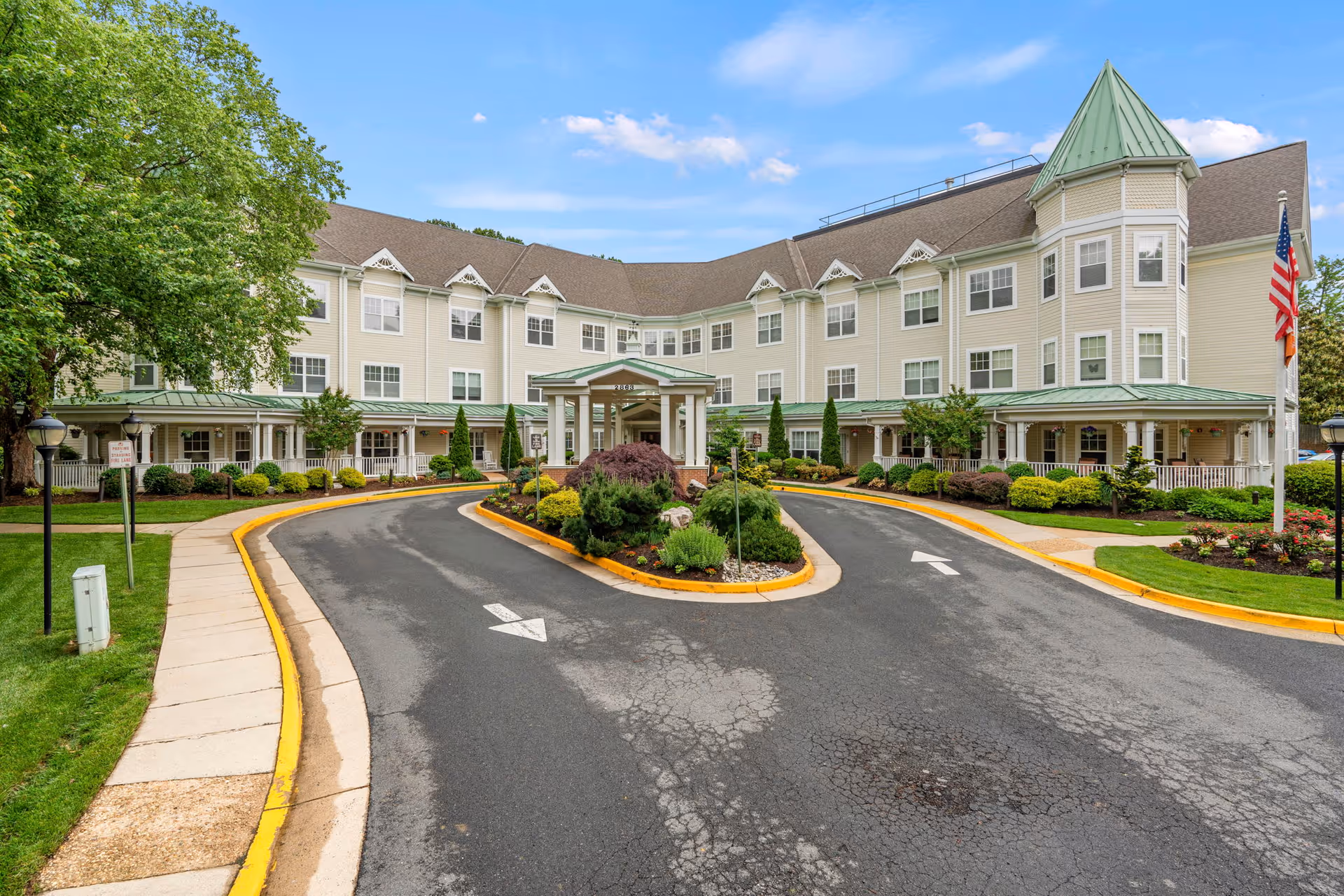 Exterior view of a large, three-story senior living facility with beige siding and green roofs. The building features a covered entrance with a circular driveway and landscaped greenery in the center. An American flag is visible on the right side, and there are trees and bushes surrounding the building under a partly cloudy blue sky.