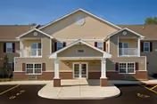 Front exterior view of a two-story residential building with beige siding, white trim, and brick accents. The building has balconies on the upper floor and a covered entrance with the number 100 above the door. There is a paved parking area in front with marked parking spaces.