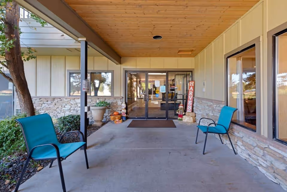 Covered outdoor entrance area of a senior living facility with two teal chairs on either side, stone wall accents, potted plants, and autumn-themed decorations including a 'HARVEST' sign and small scarecrow figures near glass double doors.