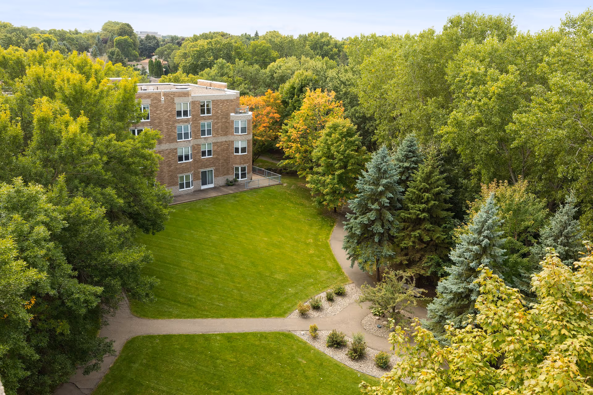 Aerial view of a senior living facility surrounded by lush green trees and well-maintained lawns. The building is a multi-story brick structure with several windows, and there are paved walking paths winding through the landscaped grounds.