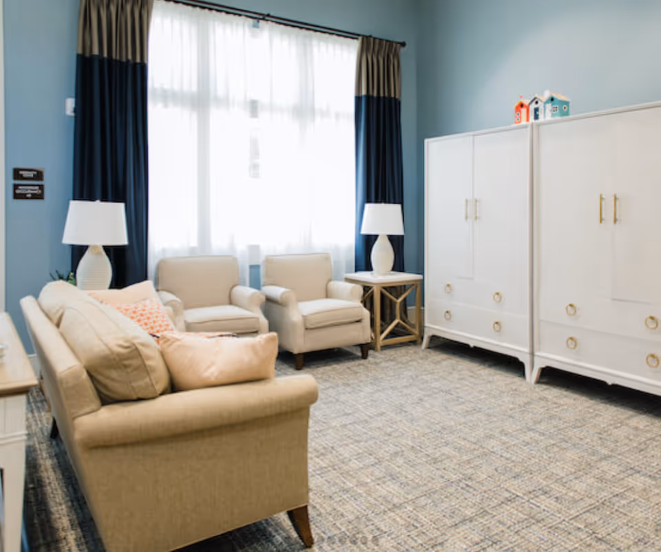 A bright sitting room with a beige sofa and two armchairs, side tables with lamps, a large window with curtains, and white storage cabinets against a blue wall.