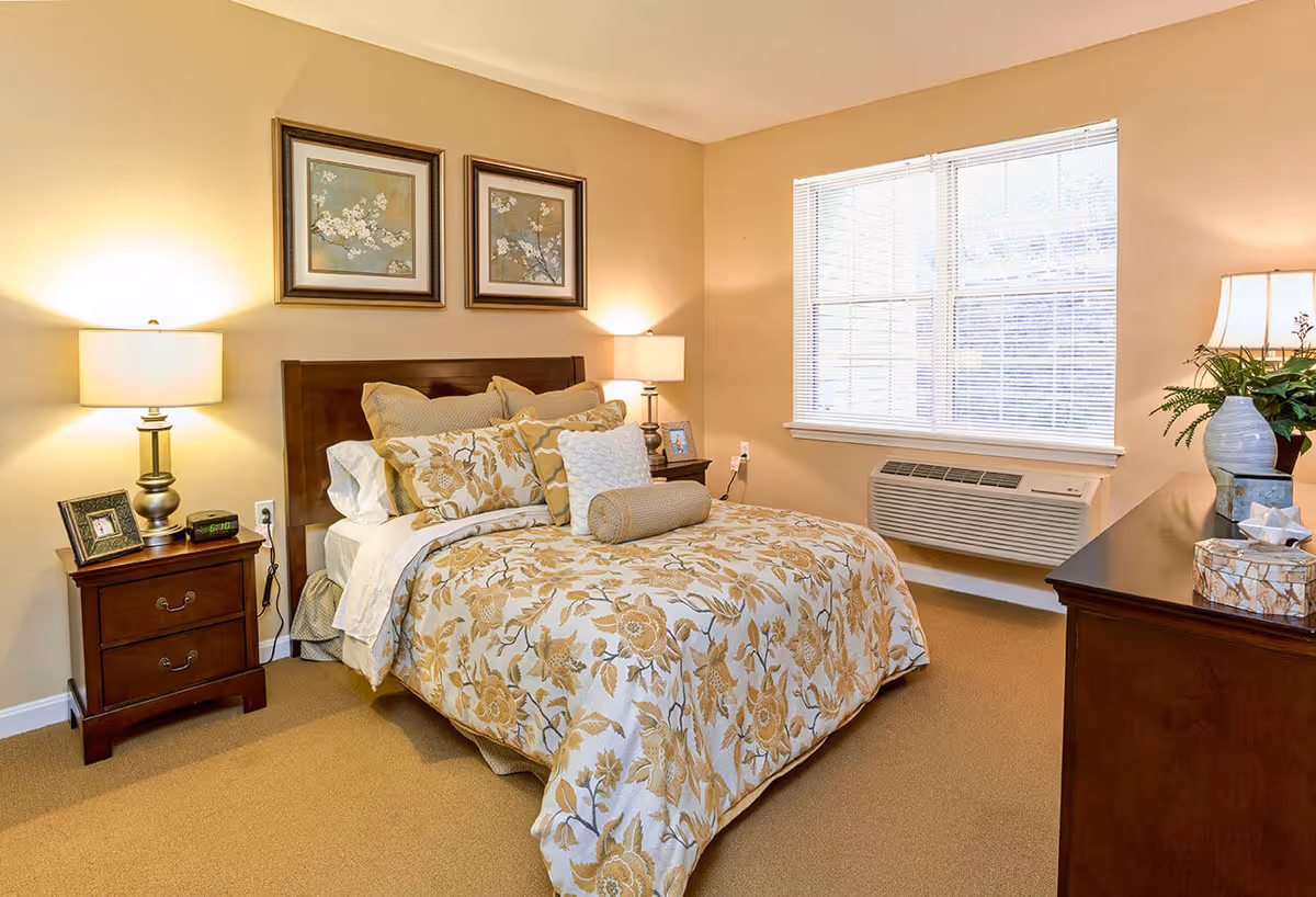 Well-lit furnished bedroom with a bed dressed in floral bedding, nightstands with lamps, a dresser, and a window air conditioner.