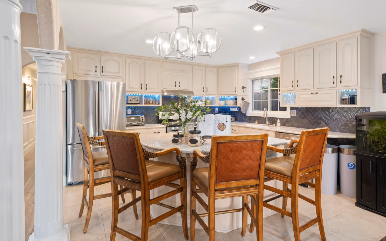 Bright and spacious kitchen with cream-colored cabinets, stainless steel refrigerator, and a large island with a granite countertop surrounded by four wooden chairs with leather seats. A modern chandelier with glass globes hangs above the island. The kitchen has a window above the sink, a dark herringbone tile backsplash, and various kitchen appliances and decor items on the counters.