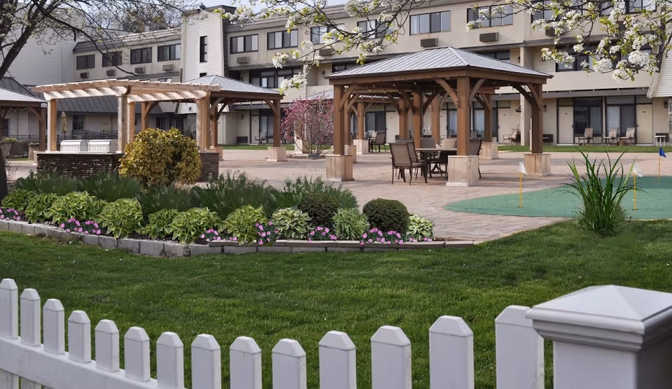 Outdoor patio area at The Arbors Assisted Living Communities at Islandia East featuring wooden pergolas with tables and chairs underneath, surrounded by landscaped greenery and flowering plants, with a white picket fence in the foreground and a multi-story building in the background.