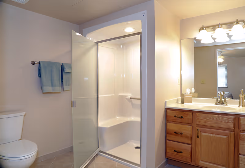 A clean and well-lit bathroom featuring a white toilet, a glass-enclosed shower with a built-in seat, a wooden vanity with a white countertop, a large mirror, and a set of blue towels hanging on a towel rack.