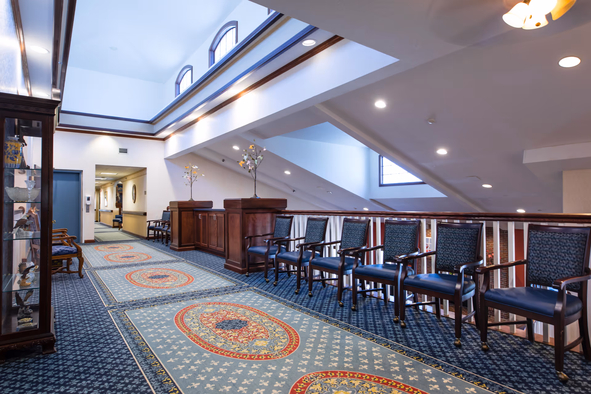 Interior common lounge hallway with a row of chairs along a railing, decorative rugs, skylights, and a display cabinet.