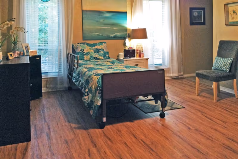 A well-lit bedroom in an assisted living facility featuring a single hospital-style bed with floral bedding, a nightstand with a lamp and decorative items, a gray upholstered chair with a patterned pillow, wooden flooring, and framed artwork on the walls.