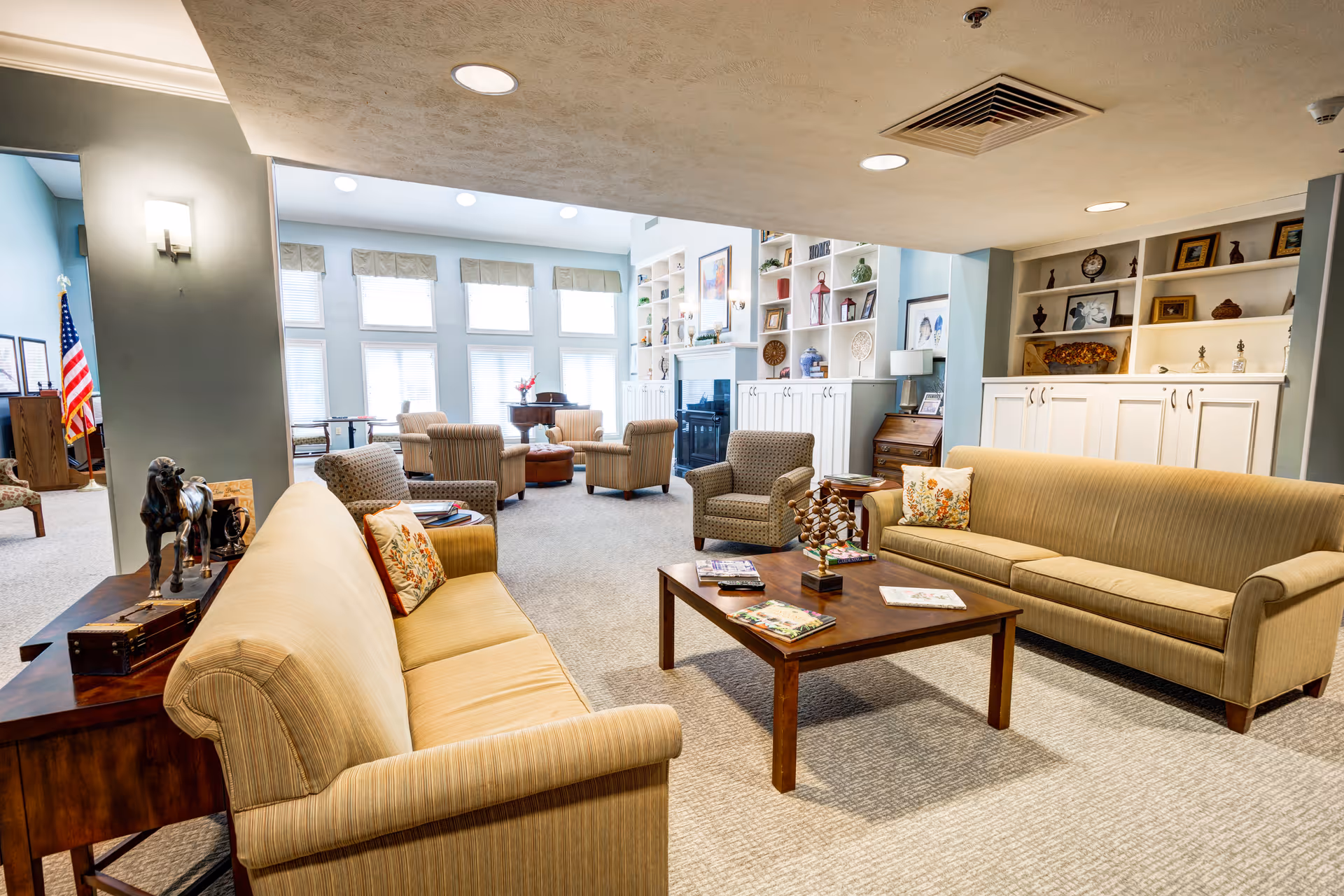 A bright and spacious living room area in a senior living facility with two beige sofas facing each other across a wooden coffee table. The room features built-in white shelves filled with decorative items, a fireplace, several armchairs, and large windows letting in natural light. An American flag is visible in the background.