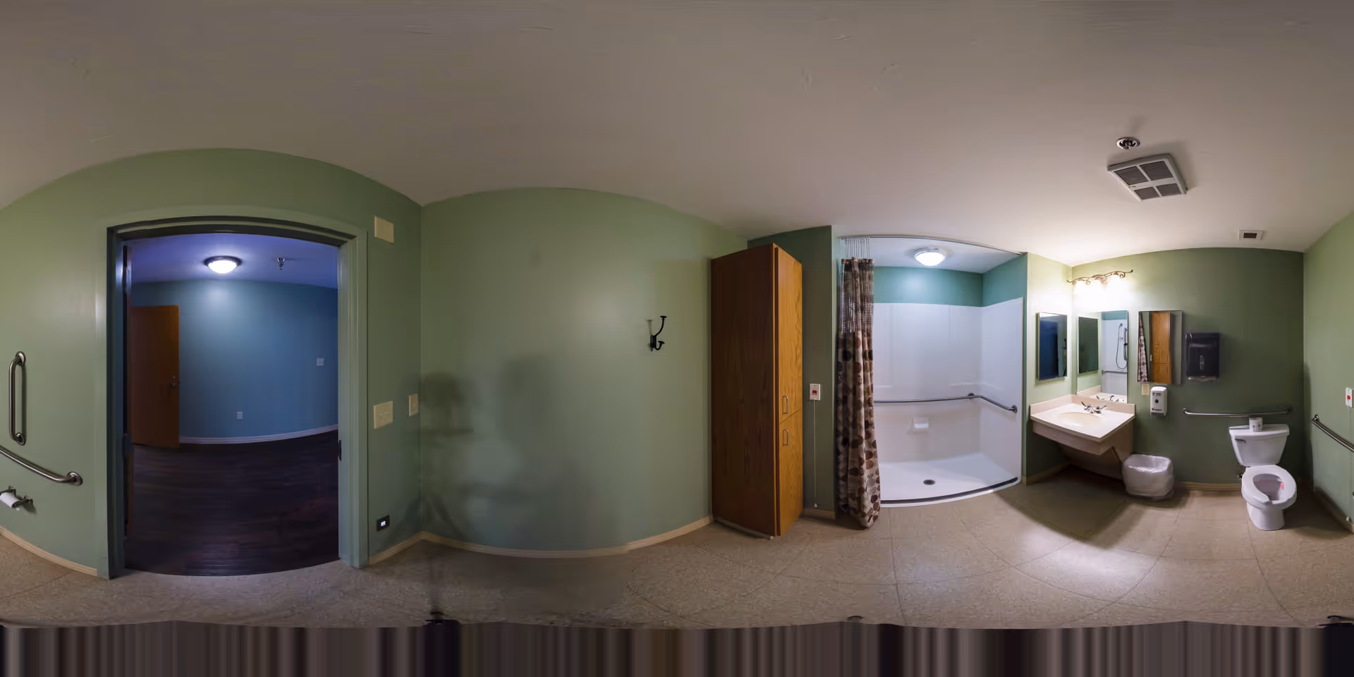 A spacious, accessible bathroom with green walls, featuring a walk-in shower with grab bars and a patterned shower curtain, a wall-mounted sink with two mirrors above it, a toilet with grab bars, a wooden storage cabinet, and a doorway leading to another room with dark flooring and blue walls.