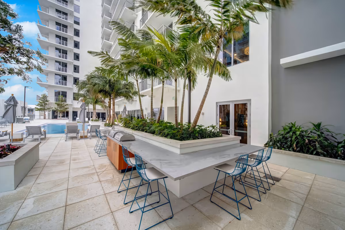 Outdoor patio area at Mirabelle featuring a modern bar-style seating with blue chairs around a large countertop planter with palm trees, adjacent to a swimming pool with lounge chairs and umbrellas, surrounded by a multi-story white building under a blue sky.