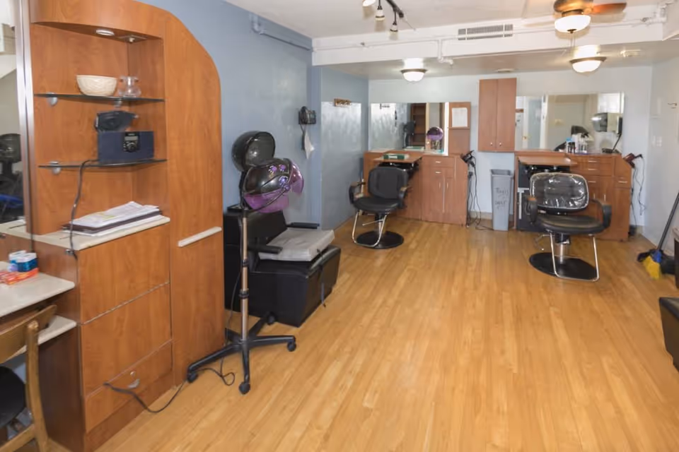 Interior salon area with hair styling chairs, hooded dryers, mirrors and wooden cabinetry on a hardwood floor.