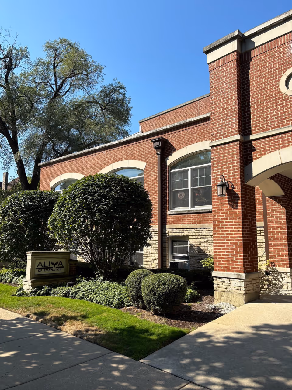 Exterior view of a brick building with arched windows and a covered entrance. There are neatly trimmed bushes and a green lawn in front of the building. A sign near the bushes reads 'ALIYA of Evanston'. The sky is clear and blue.