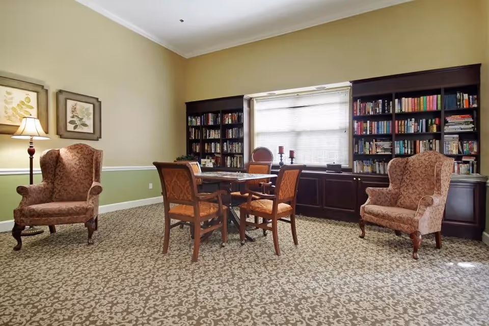 A cozy reading or sitting room with two upholstered armchairs, a wooden table with four chairs, and large built-in bookshelves filled with books. The room has patterned carpet, light green and beige walls, and a window with blinds letting in natural light.