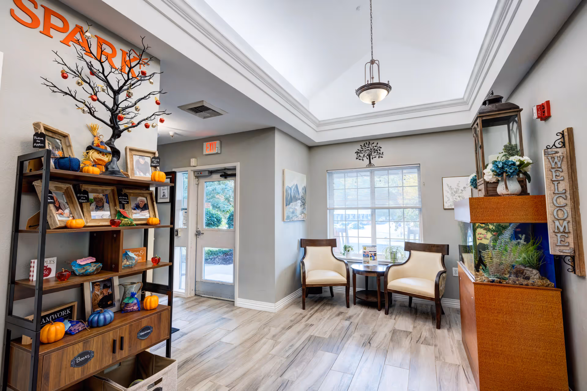 A bright and welcoming interior space with a high ceiling and a hanging light fixture. To the left, there is a wooden shelf decorated with small pumpkins, framed photos, and a black decorative tree with red and gold ornaments. Near the center, there is a glass door with an exit sign above it. To the right, two cream-colored chairs with wooden frames are positioned around a small round table in front of a window with blinds. On the far right, there is a wooden cabinet with an aquarium on top, a lantern, flower arrangements, and a vertical wooden sign that reads 'WELCOME'.