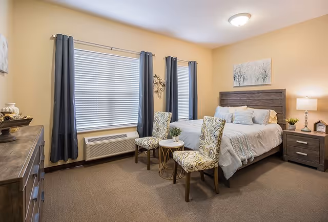 A cozy bedroom with a large bed featuring white bedding and multiple pillows. Two patterned chairs are placed in front of the bed with a small round table between them. The room has beige walls, two windows with dark curtains, a wooden dresser on the left, and a nightstand with a lamp and small decorative items on the right. A framed artwork hangs above the bed.
