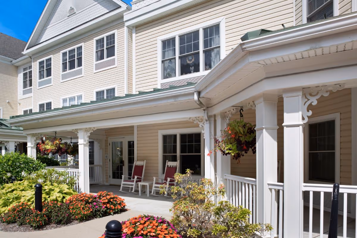 Exterior view of a senior living facility with beige siding and white trim. The building features a covered porch with white columns and decorative trim, two red rocking chairs, hanging flower baskets, and a landscaped garden with colorful flowers and greenery under a clear blue sky.