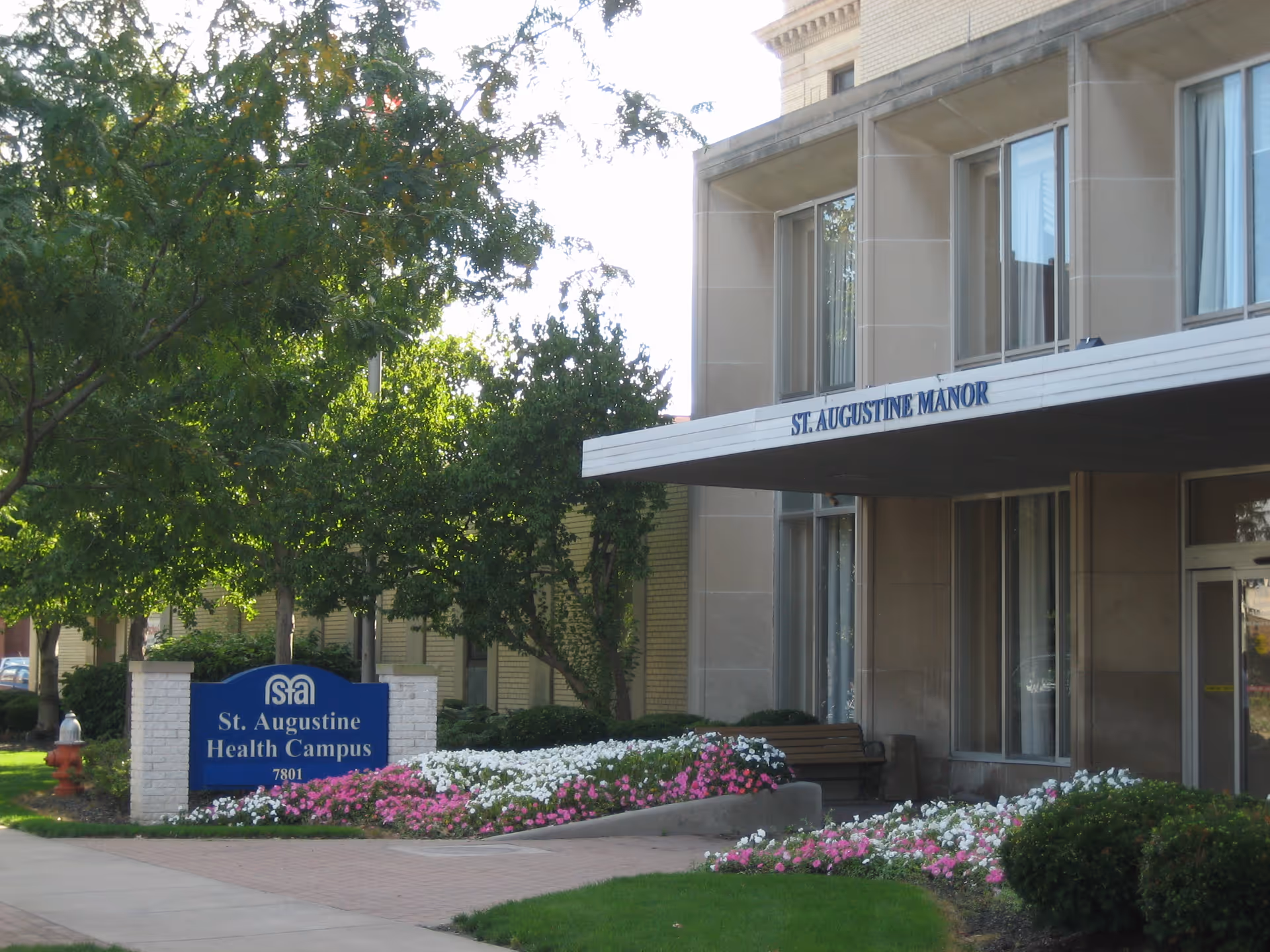 Exterior view of St. Augustine Health Campus showing the entrance to St. Augustine Manor with a covered walkway, large windows, and a landscaped area with colorful flowers and green trees. A blue sign with white text reads 'St. Augustine Health Campus 7801'.