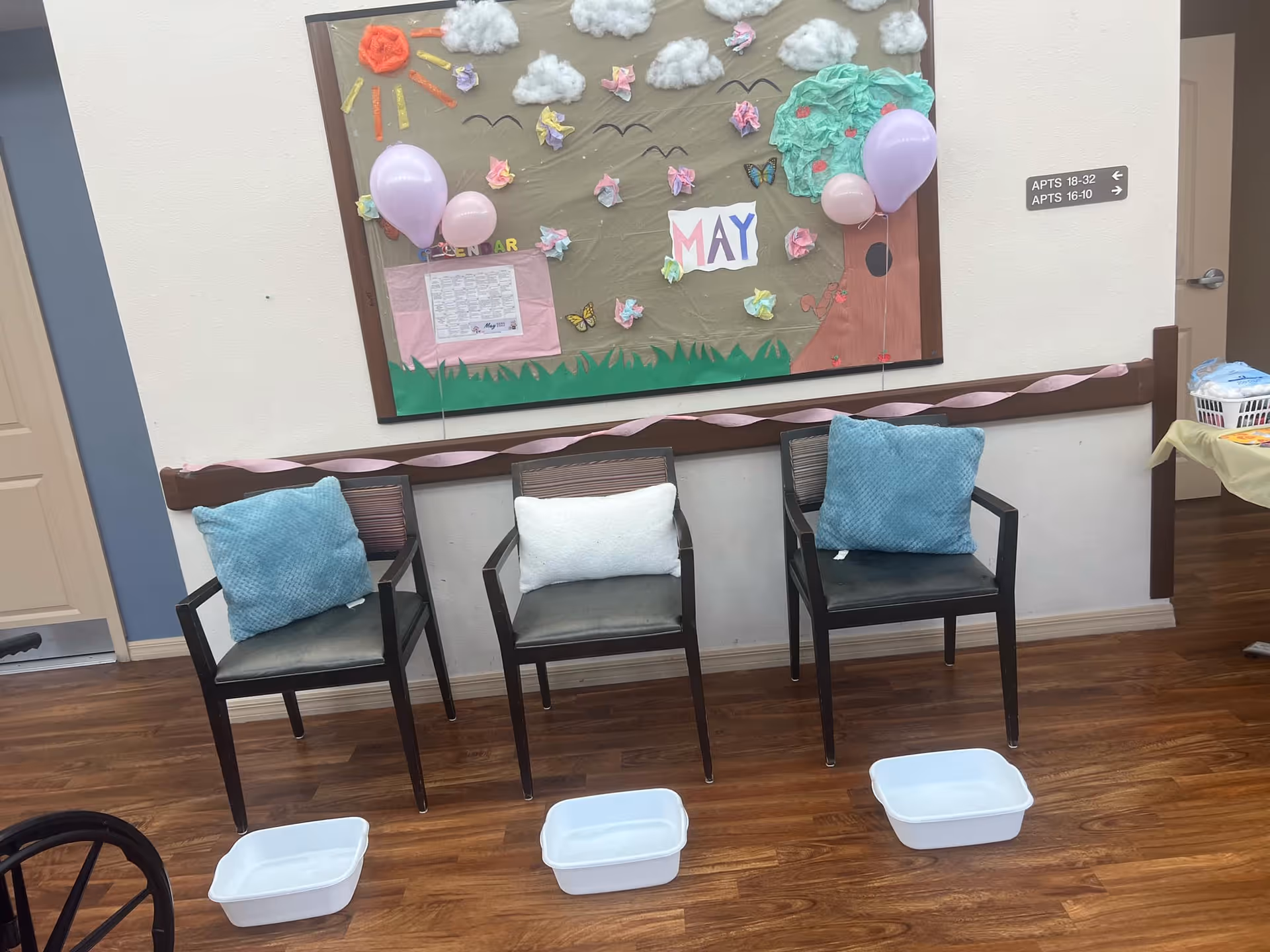 Three chairs with cushions are arranged in a row against a wall decorated with a bulletin board featuring a May calendar, paper clouds, butterflies, and balloons. Below each chair is a white plastic basin on a wooden floor. A sign on the wall indicates apartment directions.
