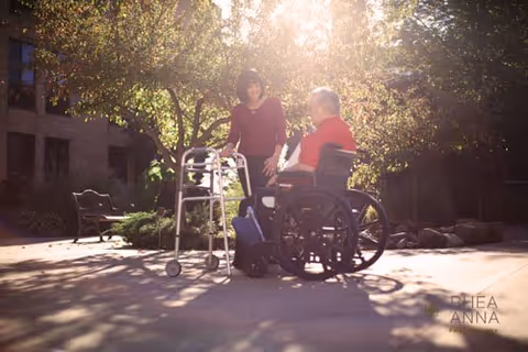 An elderly man in a wheelchair and a caregiver with a walker talking in a sunlit outdoor courtyard.