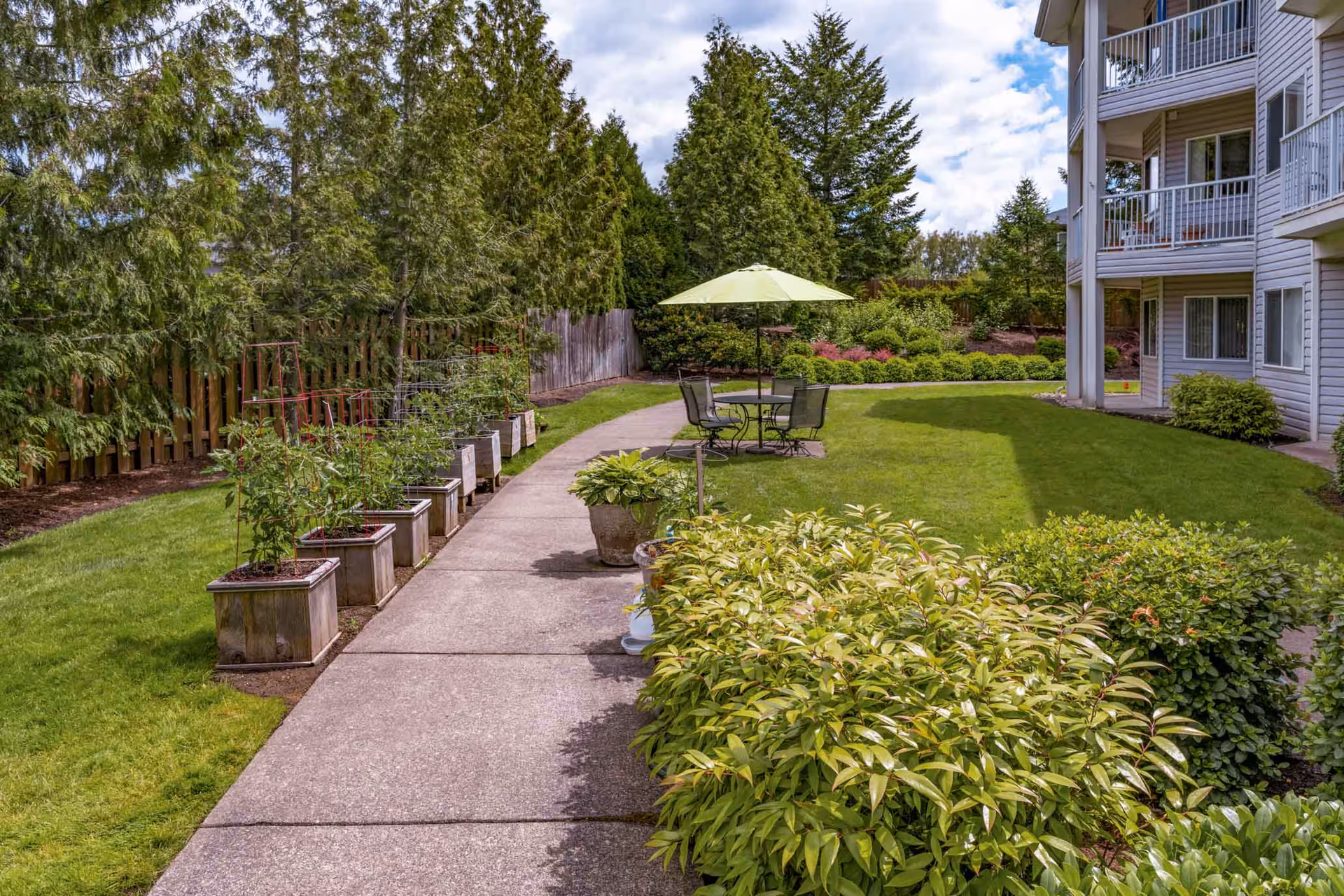 Outdoor garden area at Holiday Rock Creek featuring a concrete pathway lined with potted plants on one side and green bushes on the other. There is a round table with four chairs and a green umbrella in the grassy area. The building with balconies is visible on the right side, and tall trees and shrubs surround the garden.
