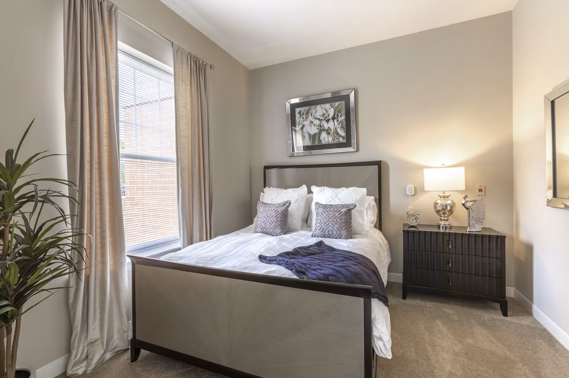A neatly made bed with white bedding and decorative pillows in a bedroom. There is a dark wooden nightstand with a silver lamp, a clock, and a decorative figurine. A large window with beige curtains is on the left side, and a framed floral artwork hangs above the bed. A tall green plant is placed near the window.