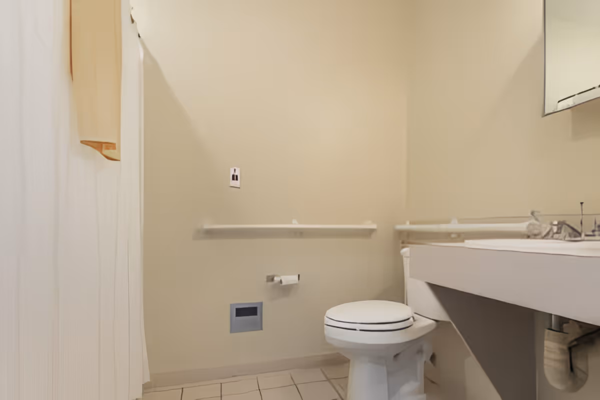 A small bathroom with beige walls and tiled floor. It features a white toilet with a closed lid, a white sink with a countertop, a mirror above the sink, and a towel hanging on the left side. There are grab bars installed on the walls near the toilet for support.