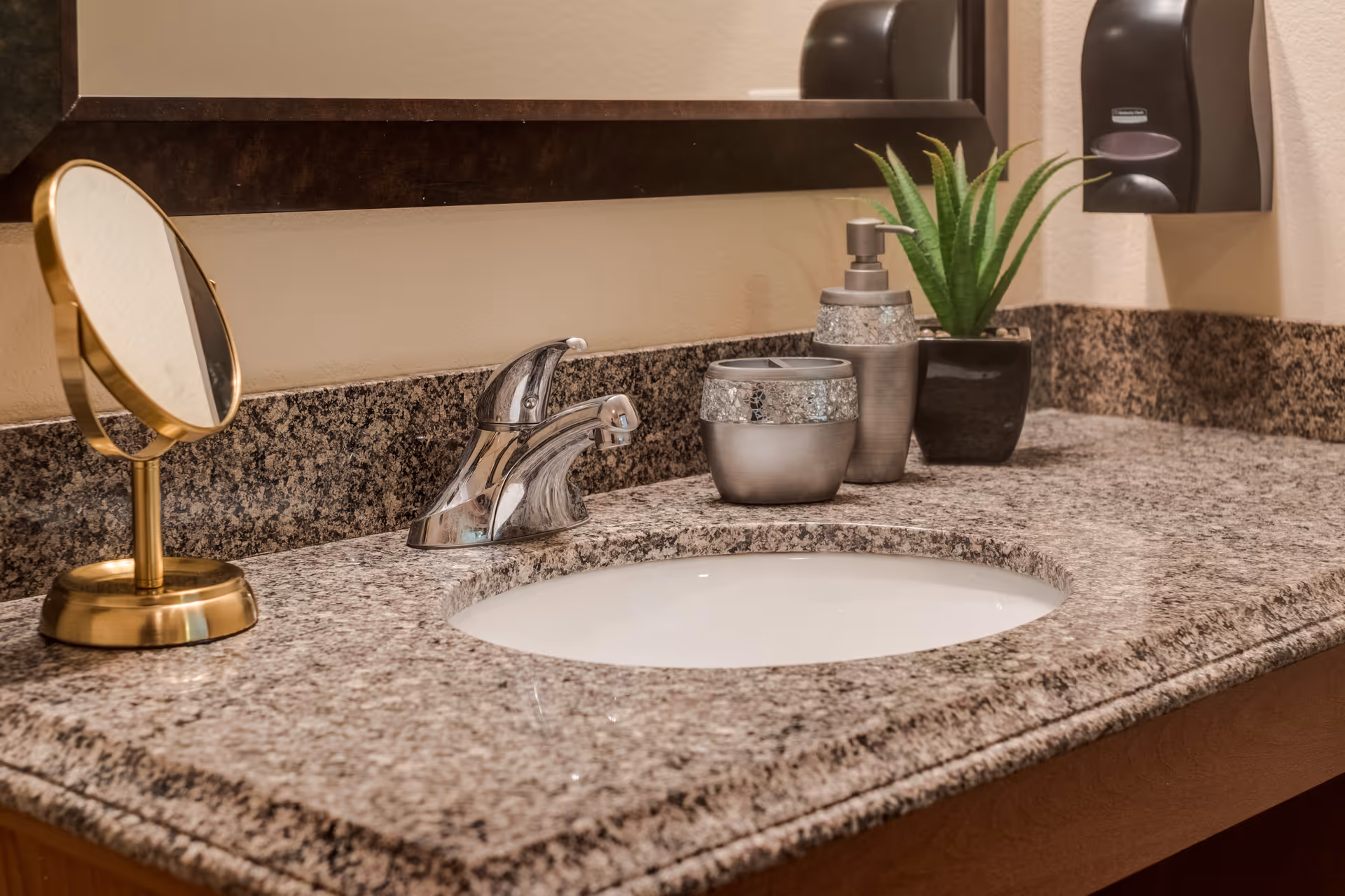 A bathroom countertop with a granite surface featuring a chrome faucet, an oval sink, a small round mirror with a gold frame, a soap dispenser, a matching container, a small potted green plant, and a wall-mounted soap dispenser.
