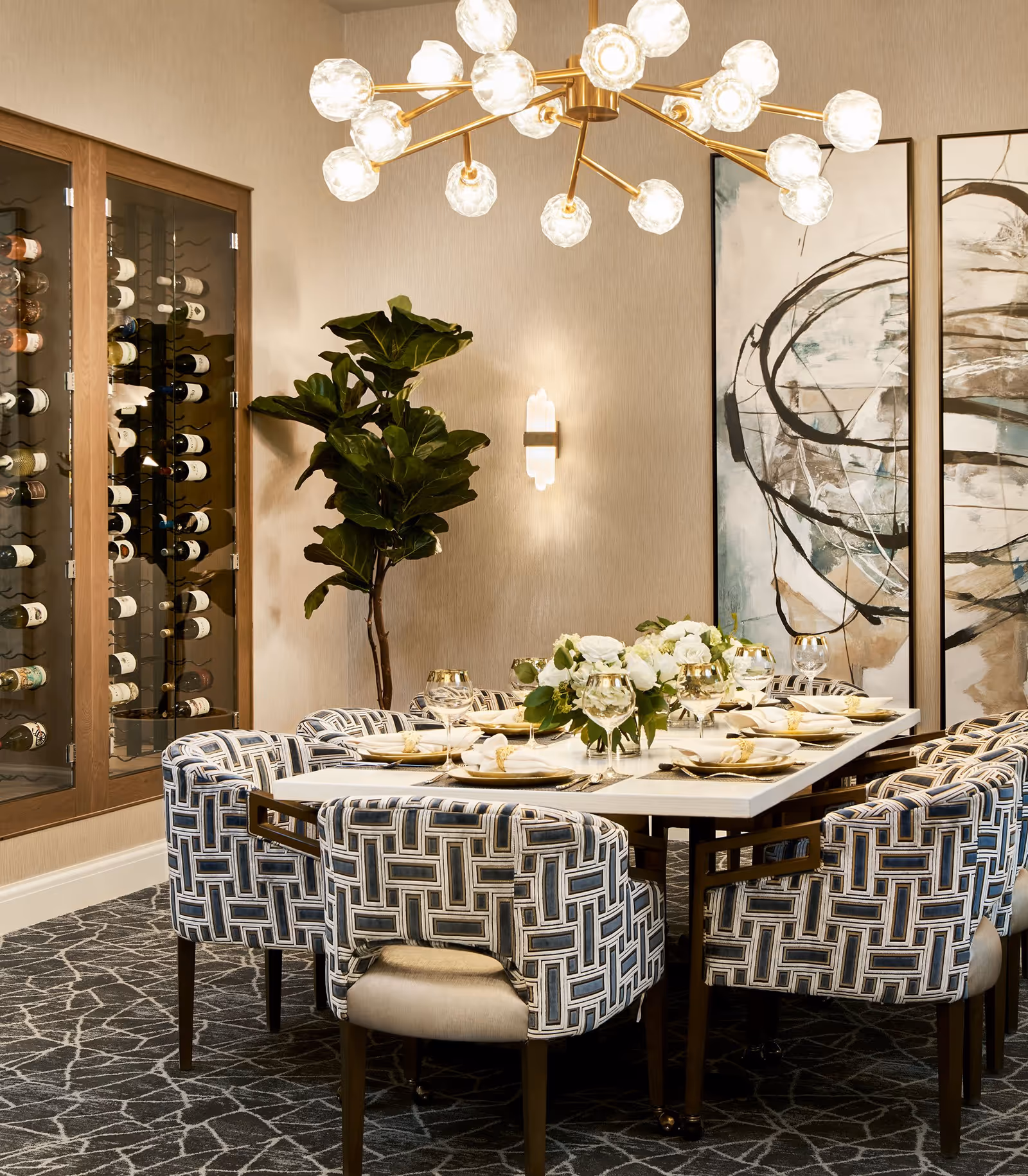 A modern dining room with a rectangular table set for eight people. The table is adorned with white plates, gold-rimmed glasses, white napkins with gold napkin rings, and a centerpiece of white flowers and greenery. Surrounding the table are upholstered chairs with a geometric pattern in shades of blue, gray, and beige. On the left wall, there is a glass-enclosed wine rack filled with bottles. A large leafy plant stands next to the wine rack. The walls are decorated with abstract artwork and a wall sconce, and a contemporary chandelier with multiple globe lights hangs above the table. The floor is covered with a patterned carpet.