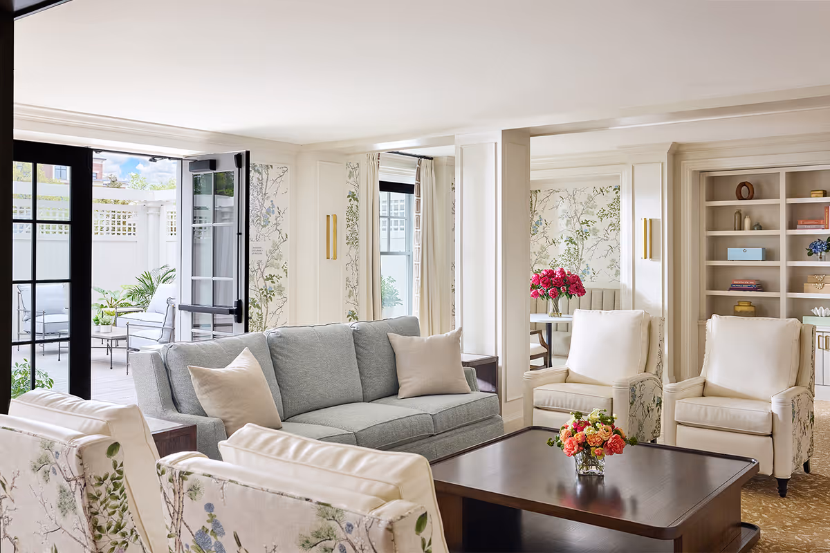 A bright and elegant living room with a light gray sofa, two cream armchairs, and two floral-patterned chairs surrounding a dark wooden coffee table with a vase of colorful flowers. The room has large windows and glass doors leading to an outdoor patio with seating. The walls are decorated with floral wallpaper and built-in shelves with decorative items.