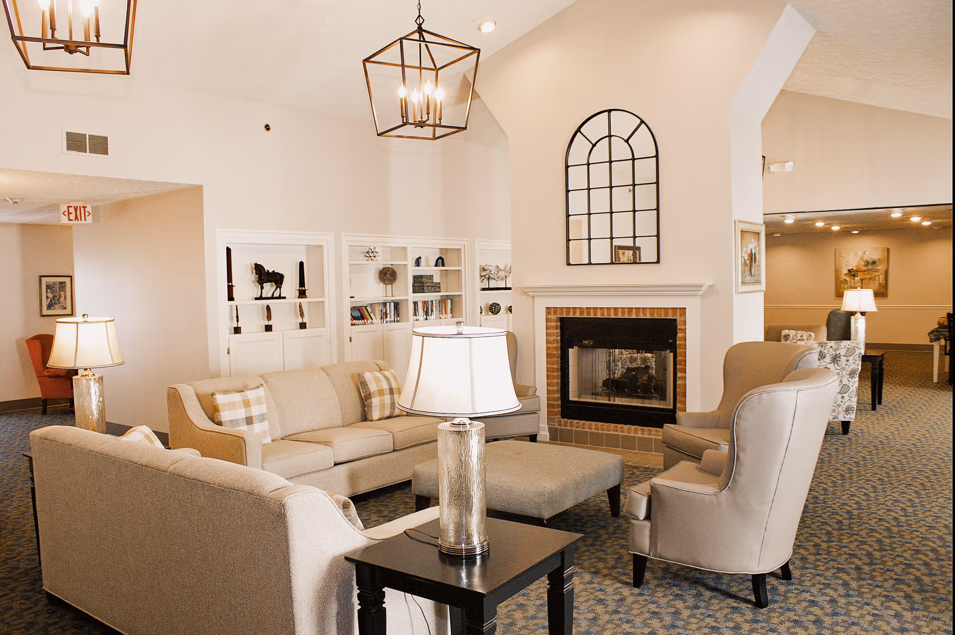 A cozy senior living community lounge area with beige sofas, armchairs, a central ottoman, and a fireplace with a decorative arched mirror above it. The room is softly lit with table lamps and hanging lantern-style light fixtures. Built-in shelves with decorative items and books are visible in the background.