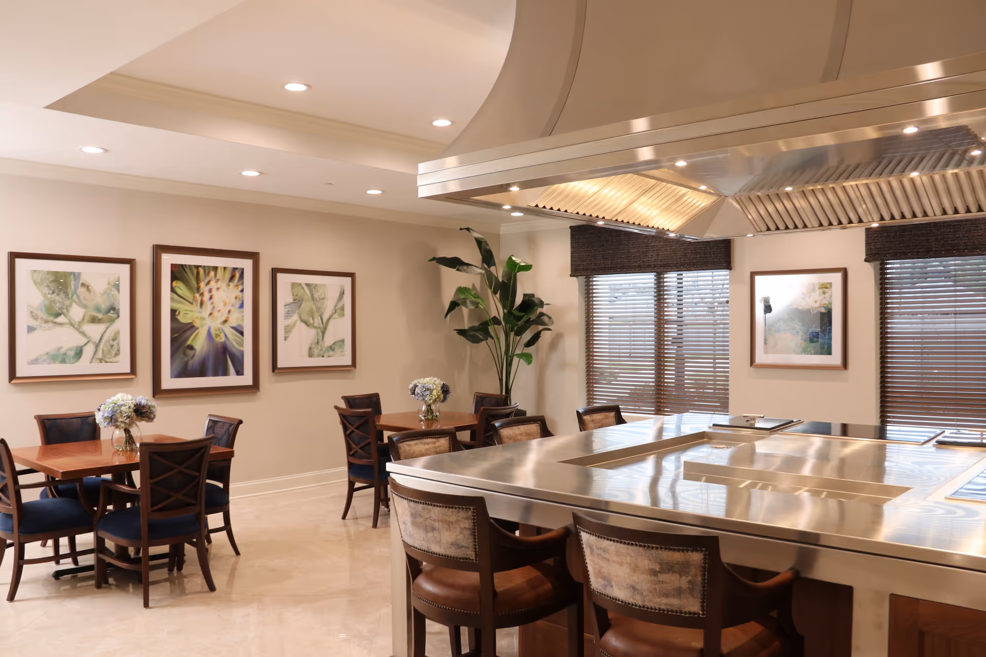 A dining area with wooden tables and chairs, each table decorated with a small vase of flowers. The room features framed botanical artwork on the walls, large windows with wooden blinds, and a large stainless steel cooking station with chairs around it. There is also a tall green plant in the corner.