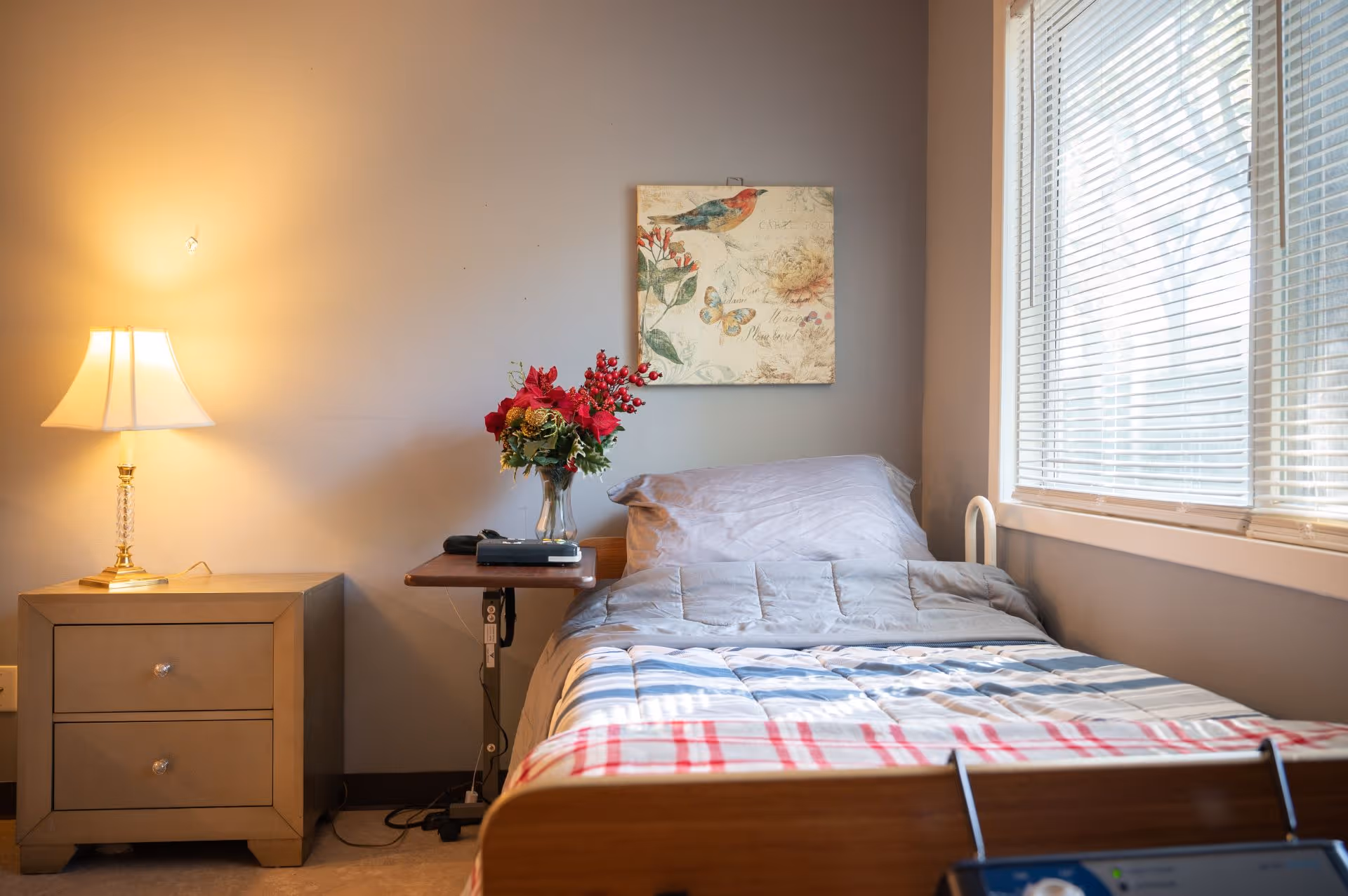 A neatly made single bed with a gray comforter and a red and white striped blanket at the foot. Next to the bed is a wooden nightstand with a lamp and a small table holding a vase of red flowers and a remote control. A window with blinds is on the right side, and a wall painting featuring a bird and flowers hangs above the bed.